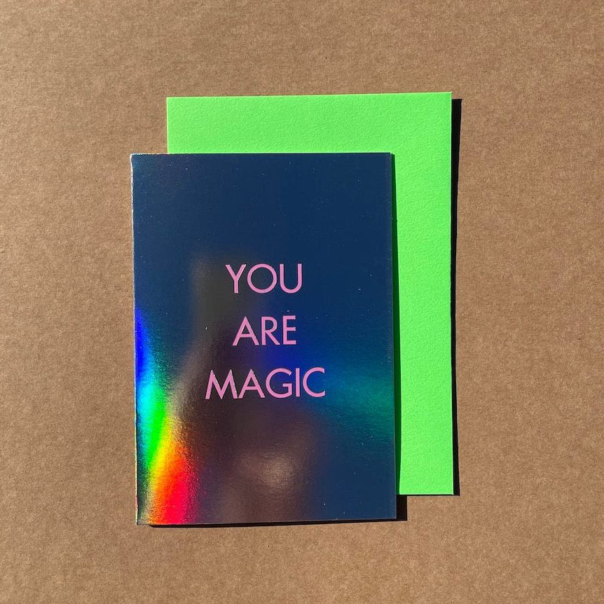 A greeting card with holographic foil and pink text reading 'YOU ARE MAGIC', placed on top of a bright green envelope against a brown kraft paper background.