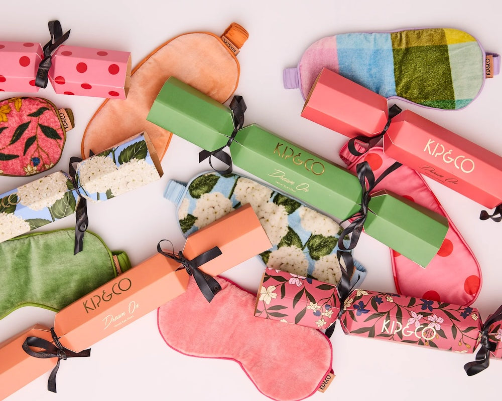 A styled flat-lay of KIP&CO home spa products on a white background, featuring many colourful sleep masks, and their matching Christmas bon-bon-shaped gift boxes tied with black ribbons.