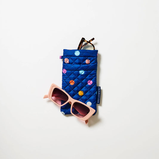 Styled image of Blue quilted sunglasses case with colorful polka dots and pink sunglasses and a pair of reading glasses peeking from the top of case on a white background