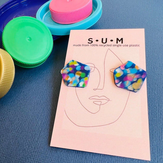 A pair of confetti blue colored stud earrings made from recycled plastic, displayed on a card with the brand name 'S U M' at the top, along with various other colors of recycled plastic lids in the background.