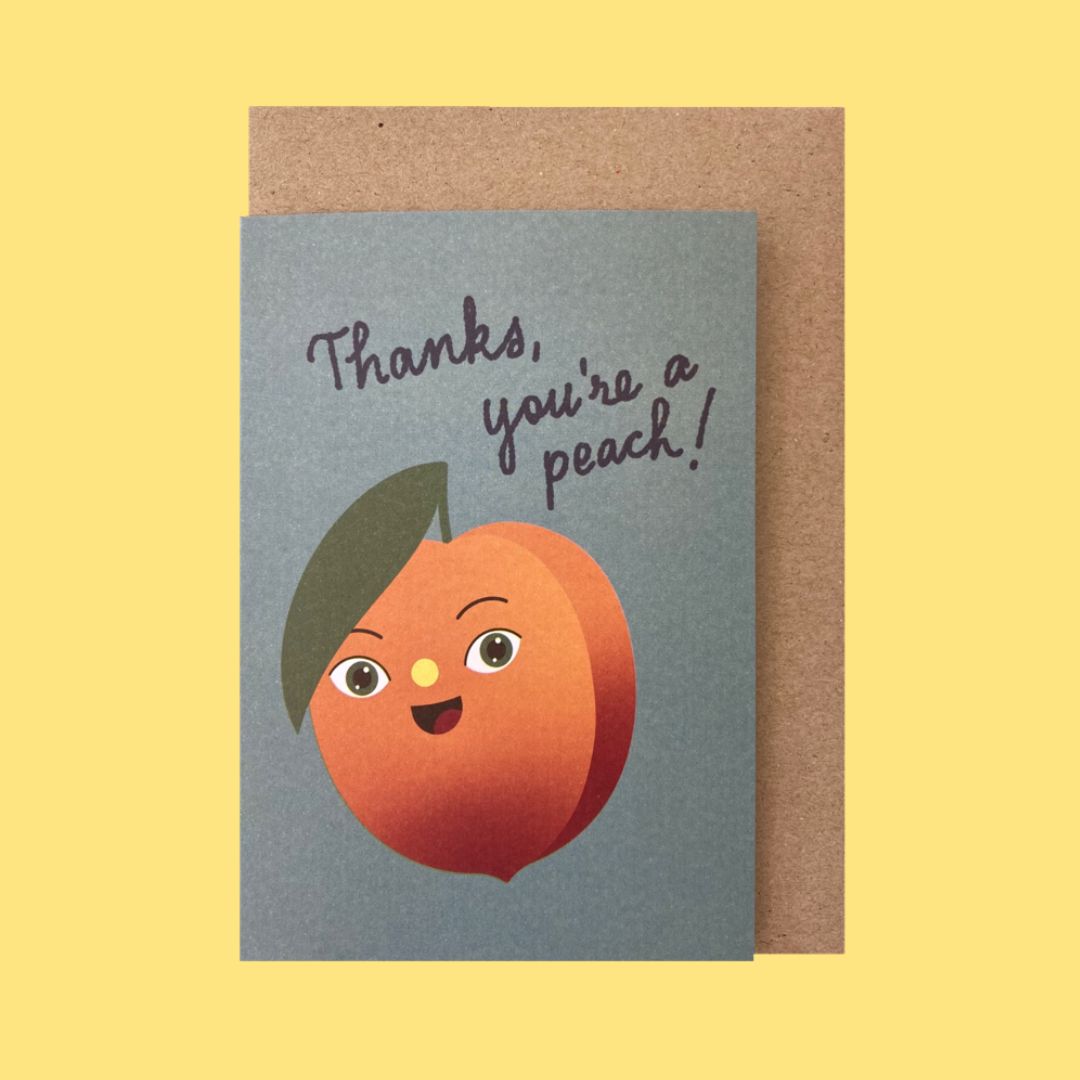 A greeting card with a blue-gray background featuring a cute illustrated peach with a leaf and the text 'Thanks, you're a peach!' in dark script, shown with a brown kraft envelope on a pale yellow background.