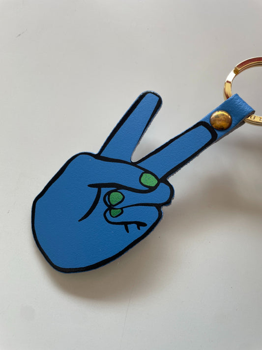 A blue leather keychain shaped like a hand making a peace sign, with green painted fingernails, black outlines, and a gold-tone key ring.