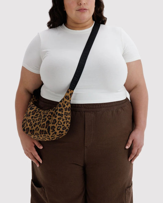 A model wearing a leopard print crossbody bag with a black strap, styled with a white t-shirt and brown pants against a white background.