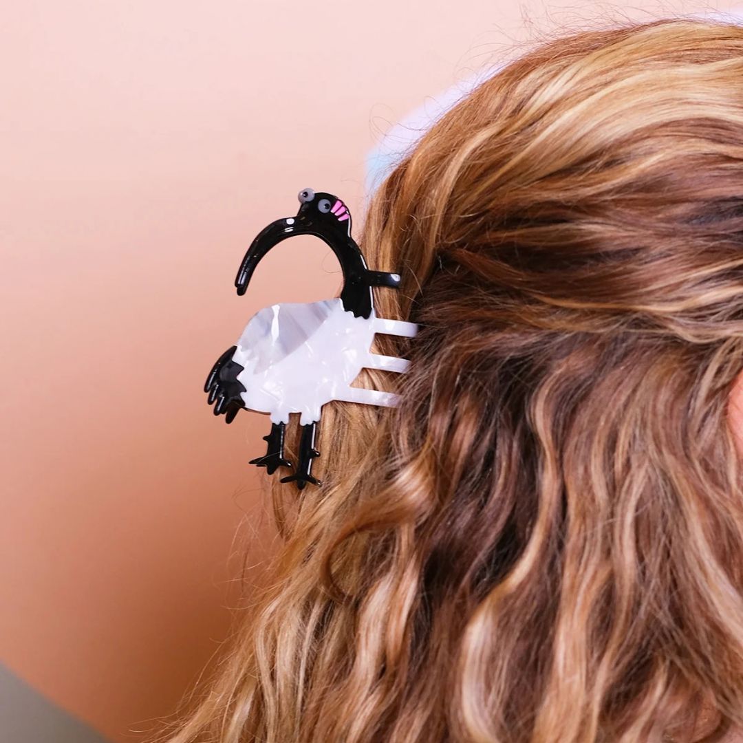 A close-up lifestyle shot of a black and white Ibis hair clip holding back a person's wavy blonde hair in the half 'n' half style, against a solid peach background.