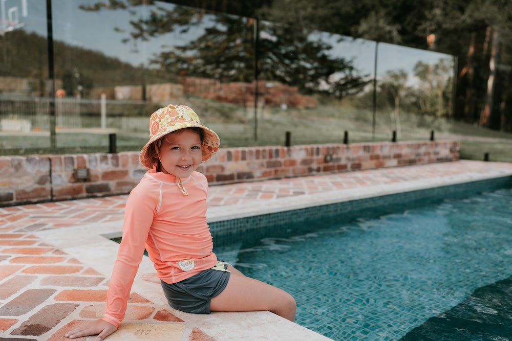 front view of Bedhead Tallulah Swim Bucket Hat worn by a young model sitting on the edge of a swimming pool