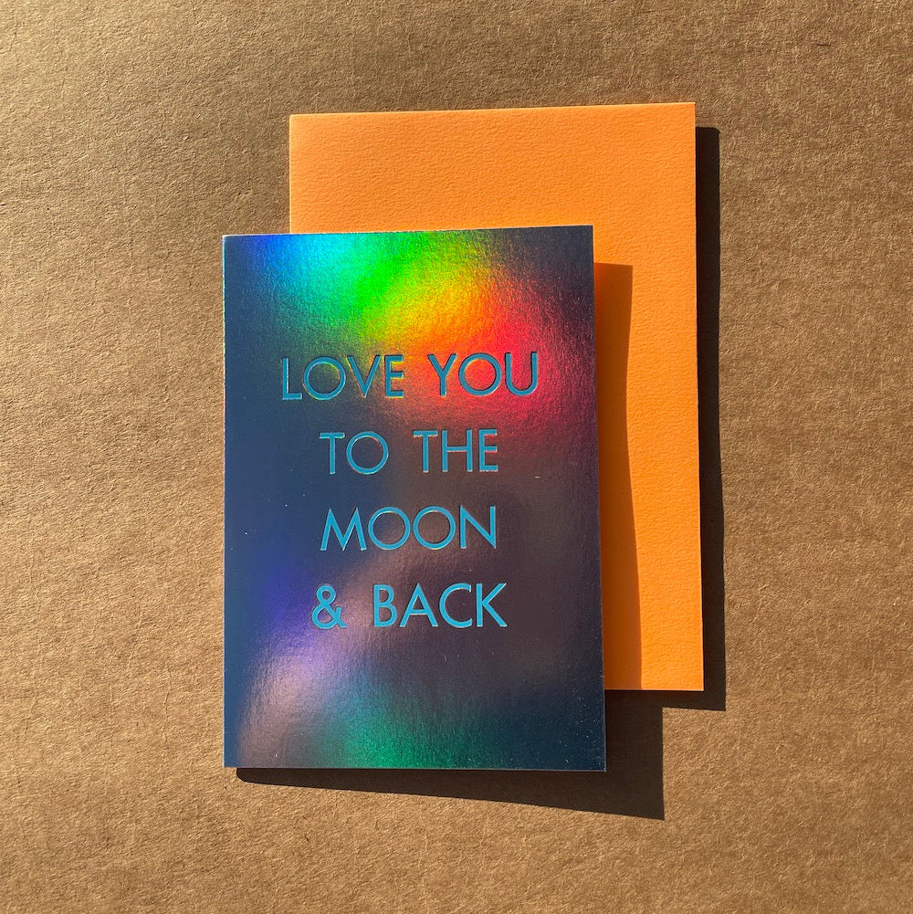 A greeting card with an iridescent holographic background and the phrase 'LOVE YOU TO THE MOON & BACK' in light blue text, shown with an orange envelope on a brown textured surface.