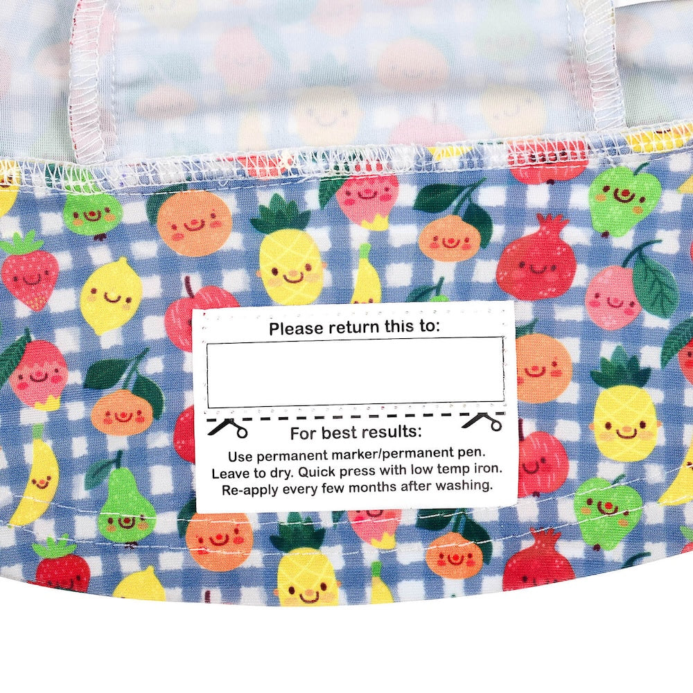 Close-up of the name label on the inside brim of a bucket hat, featuring a colorful print of smiling fruits like pineapples, apples and oranges on a blue gingham background.