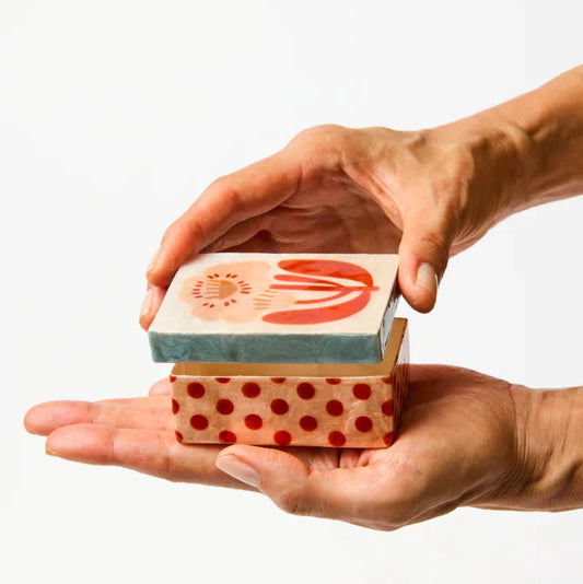 Two hands holding a small shell box with a flower and polka dot motif