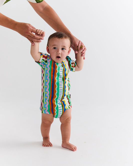Baby in a colorful striped onesie standing with assistance on a white background