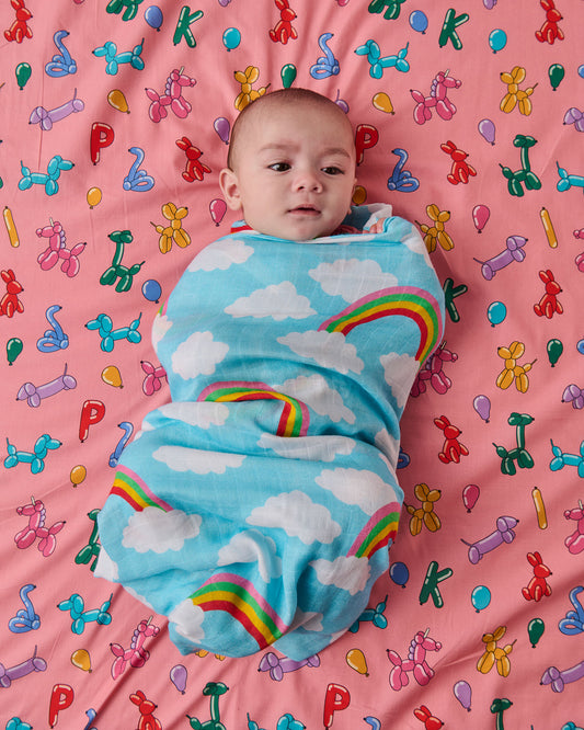 Baby swaddled in a blue blanket with clouds and rainbows on a pink background with colorful balloon designs.