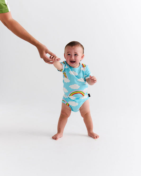 Baby in a blue onesie with rainbow and cloud patterns, standing on a white background.