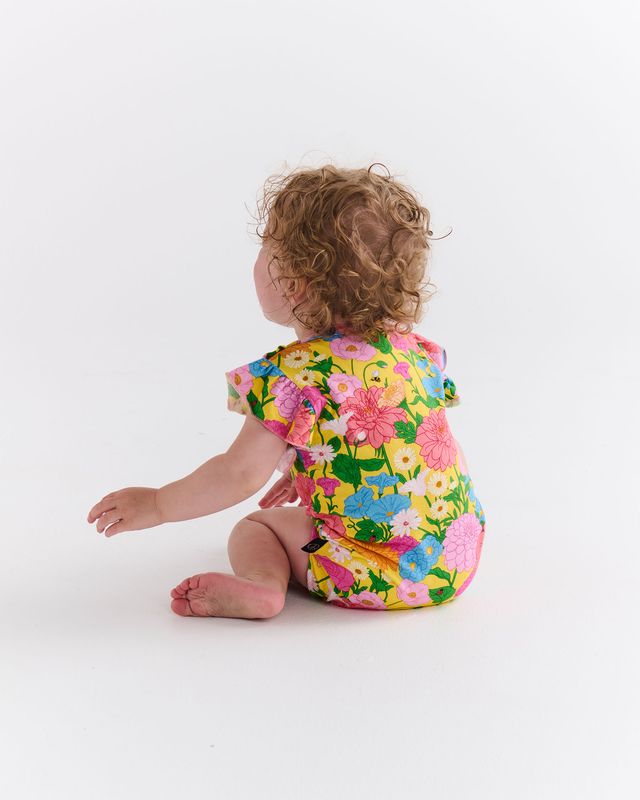 Child sitting with their back towards the viewer, wearing a colorful floral romper, on a white background