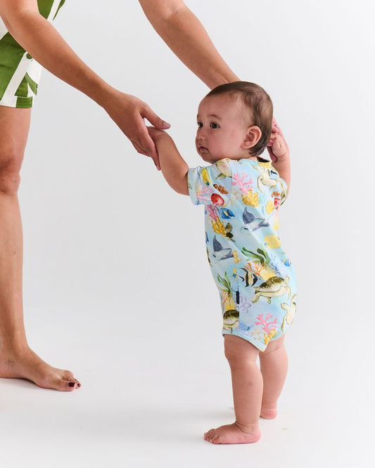 Baby in a colorful sea-themed onesie standing with an adult's hand on a white background