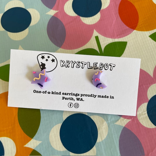 Pair of star and moon shaped earrings in purple with pink and yellow squiggles, on a KRYSTLEBOT branded card