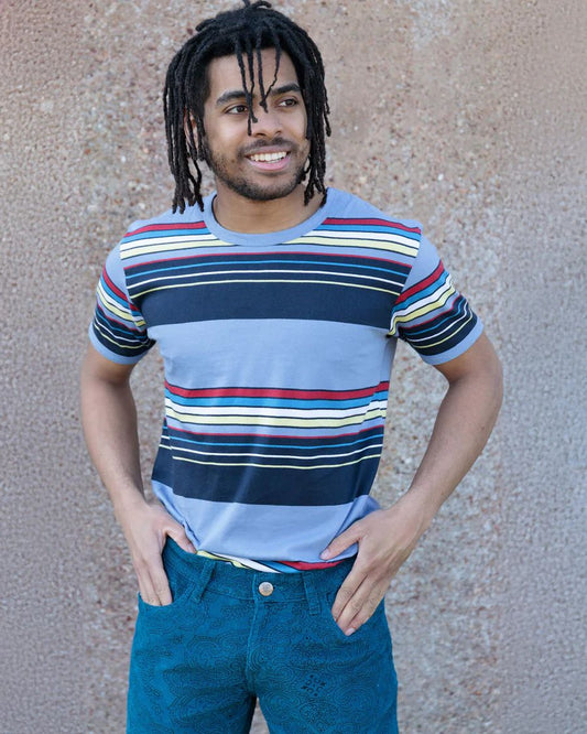 Model is wearing Run and Fly Blue Stripe Tee and smiling and looking away from the camera with both hands in pant pockets