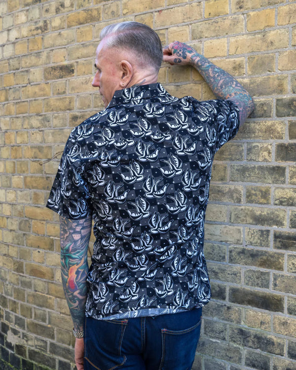 Back view of model wearing the Run and Fly Swallows Checkerboard Shirt. Model is leaning against the brick wall and looking away from the wall.