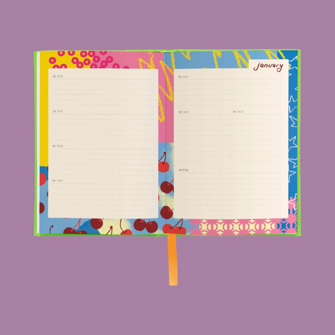An open notebook or planner with lined pages and a colorful patterned cover, shown on a solid lavender background. The cover features cherry illustrations and abstract shapes in pink, blue, yellow, and green, with an orange bookmark ribbon hanging from the bottom.