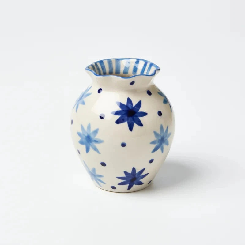 A small ceramic vase with a cream-colored body, decorated with blue floral starburst motifs, blue polka dots, and blue-and-white striped detailing on the rim, set against a plain white background.