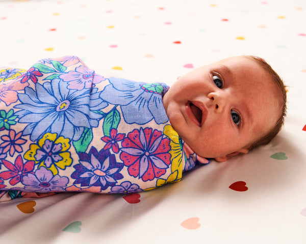 Styled image - side view of a baby swaddled in Kip & Co Bunch of Fun Bamboo Baby Swaddle on a bed with hearts on the bedsheet.