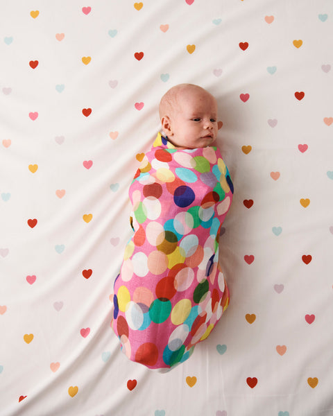 Styled image - Bird's eye view of Kip & Co Confetti Fun Bamboo Baby Swaddle wrapped around a baby on a bed with hearts on the bedsheet.