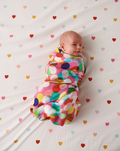 Styled image - Bird's eye view of Kip & Co Confetti Fun Bamboo Baby Swaddle swaddled around a baby on a bed with hearts on the bedsheet.