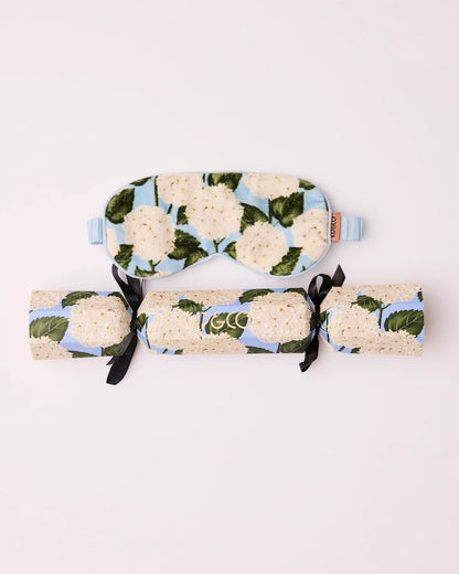 A velvet sleep mask and its matching packaging that looks like a Christmas bon-bon with a light blue and cream hydrangea floral print, tied with black ribbons, on a plain white background.