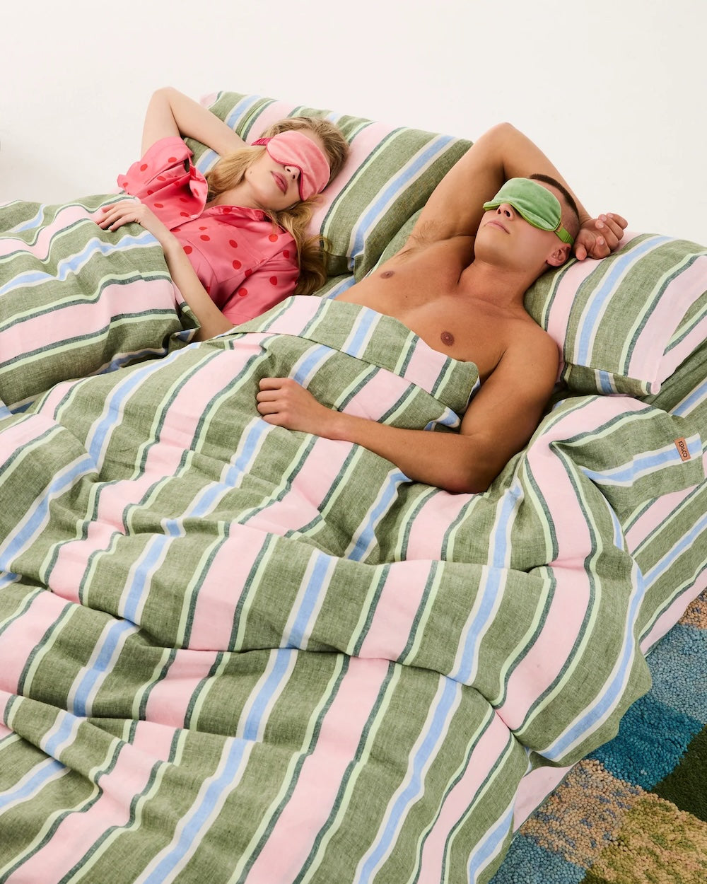 Styled image of a man and a woman laying in a bed under a striped blanket and pillow set. The woman is on the left  and is wearing a pink velvet eye mask over her eyes, and a pink and red polka dot top. The man is on the right wearing a green velvet eye mask over his eyes,  and he is not wearing a top and has one of his arms over his head.