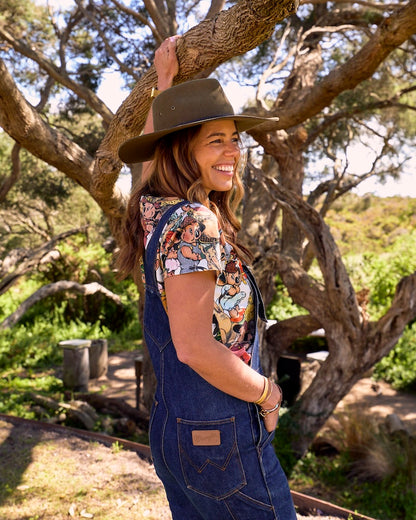 A woman outdoors under a tree wearing a wide-brimmed brown hat, a colorful Blinky Bill printed short-sleeve patterned top, and blue denim overalls. She is smiling and looking over her shoulder in a natural outdoor setting.