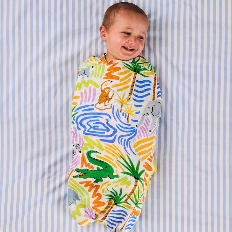 A baby smiling while wrapped in a colorful safari-themed swaddle blanket with palm trees, crocodiles, elephants, monkeys, and zebras, lying on a blue and white striped sheet.