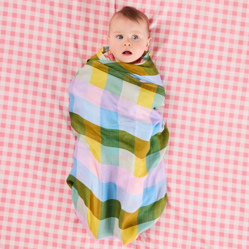 A baby wrapped in a pastel plaid swaddle blanket, lying on a pink and white gingham blanket. The swaddle features soft blocks of green, yellow, light blue, and lavender.