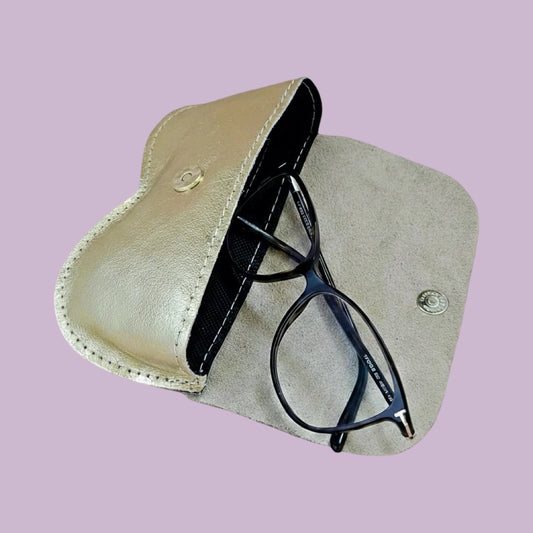 A pair of black metal-framed eyeglasses resting inside an open light beige leather glasses case with a snap-button closure, set against a solid lavender background.