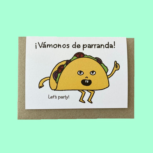 A white greeting card with a brown kraft envelope, featuring a cartoon taco character and the Spanish text '¡Vámonos de parranda!' with 'Let's party!' below, set against a solid mint green background.