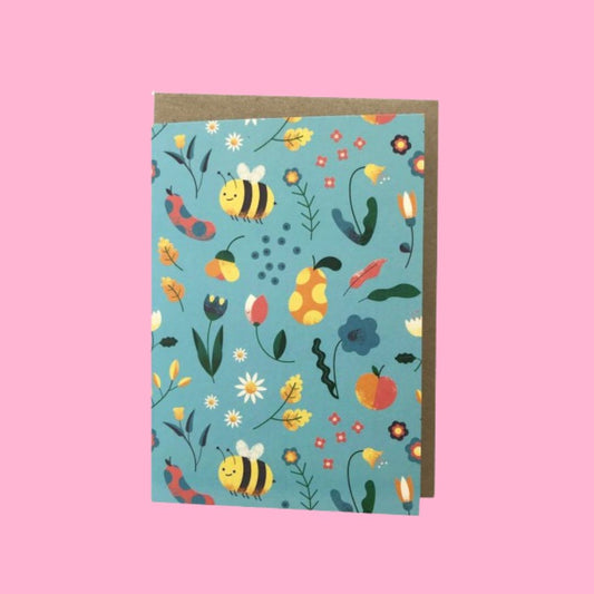 A greeting card with a colorful nature-themed pattern of bees, flowers, leaves, and acorns on a light blue background, shown with a brown kraft envelope against a solid pink backdrop.