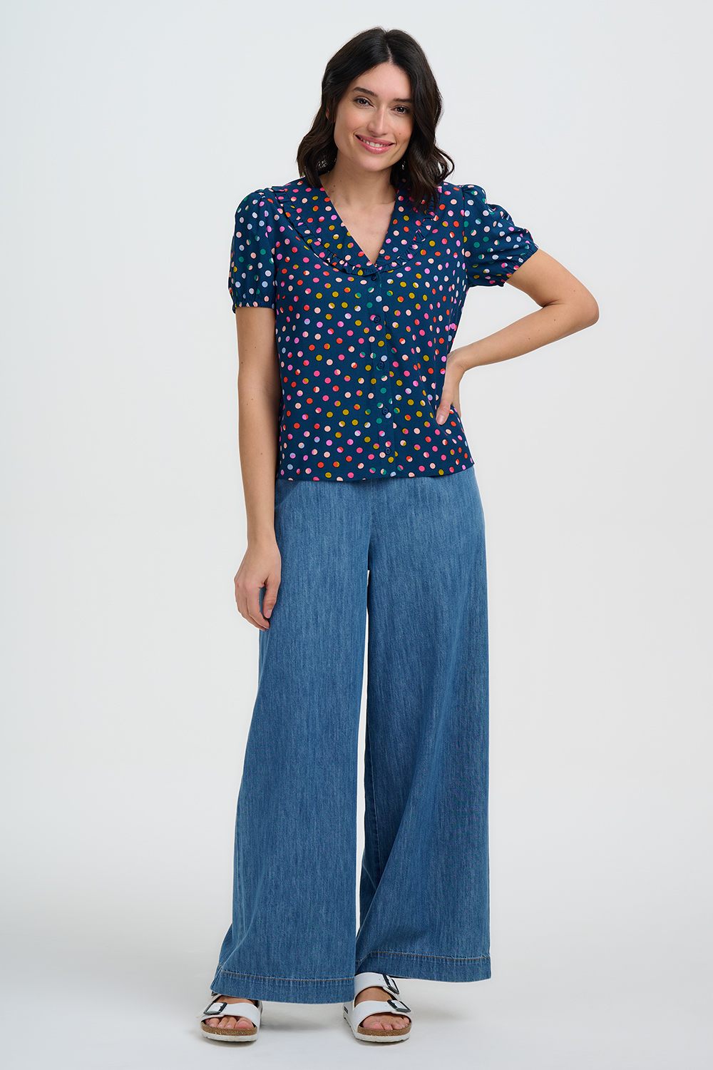 Woman wearing a navy polka dot top and wide-leg blue pants with one arm on hip on a white background