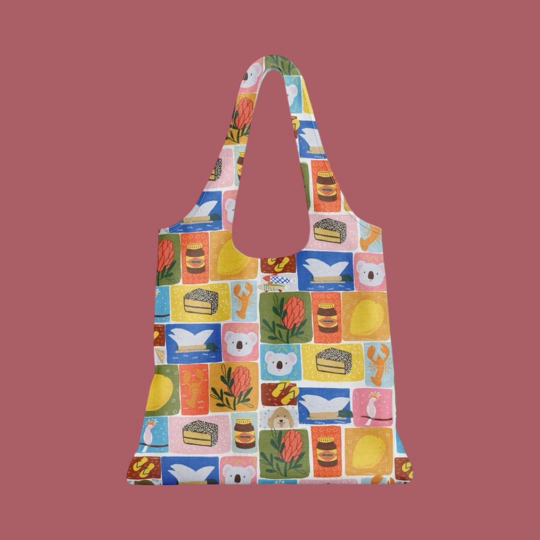 A reusable tote bag with a colorful patchwork print of illustrated Aussie icons including food items, flowers, and other icons, shown against a solid dusty rose background.