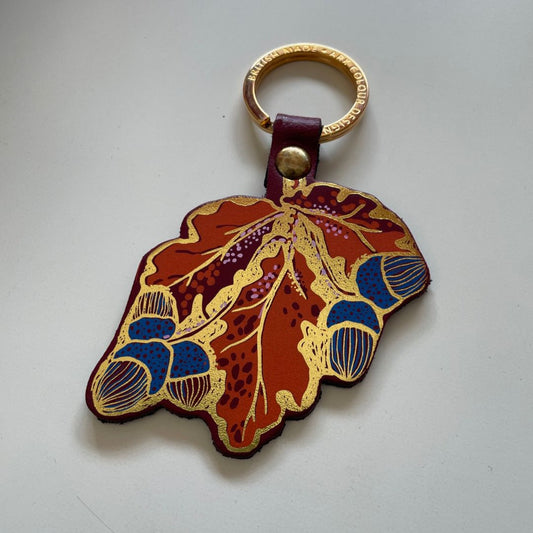 A leather keychain featuring acorns and leaves and outlined in gold foil. 