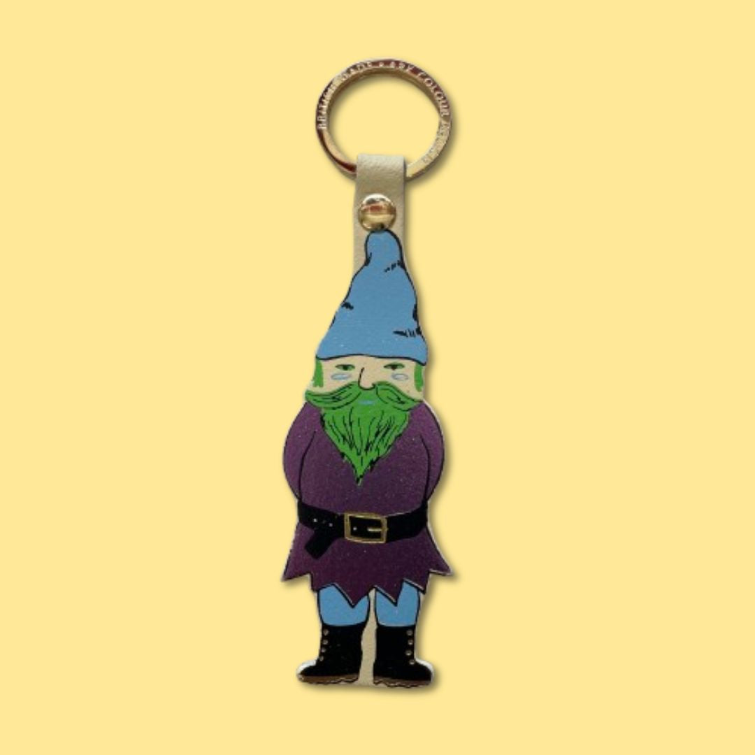 A small gnome keychain charm with a blue hat, green beard, metallic purple outfit, and black boots gold accents, attached to a metal key ring against a pale yellow background.