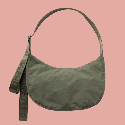 Baggu Medium Crescent Bag in Caper. An olive green crescent-shaped shoulder bag with a matching wide strap, shown against a solid pink background. The brand name 'BAGGU' is embossed on the strap.
