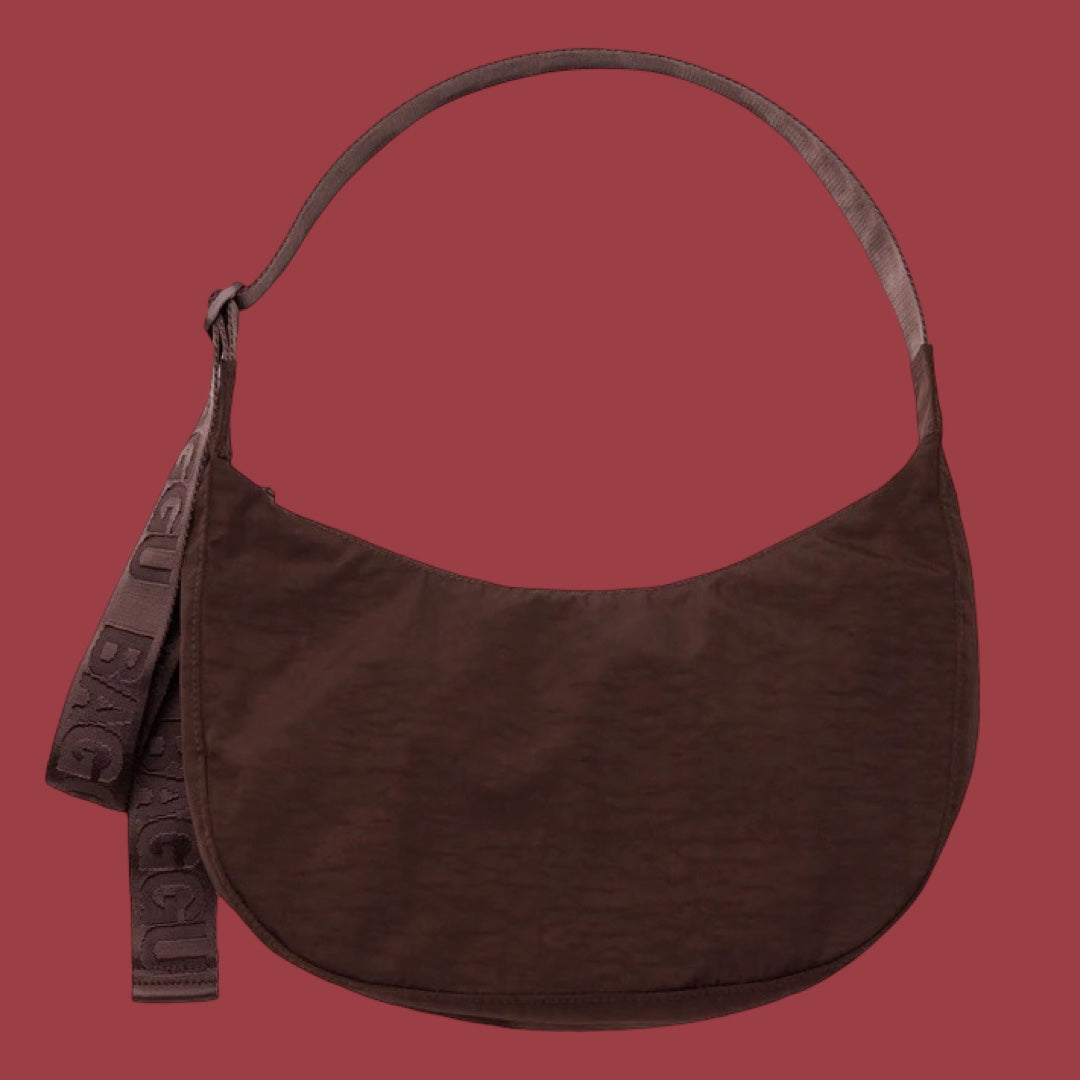 Baggu Medium Crescent Bag in Coffee. A dark brown crescent-shaped shoulder bag with a matching strap, shown against a solid dark red background. A hanging tag on the left side features the brand name 'BAGGU' in embossed lettering.