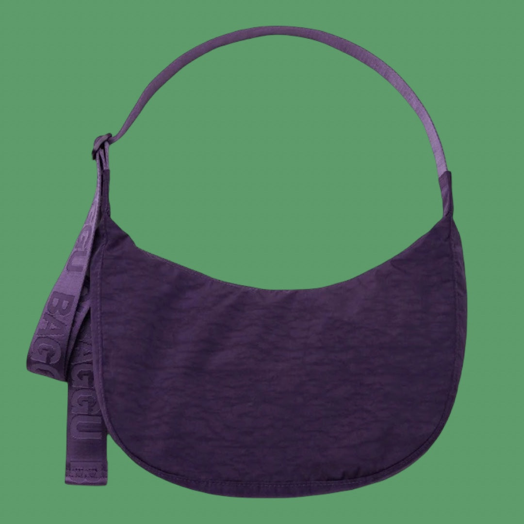 Baggu Medium Crescent Bag in Fig. A dark purple crescent-shaped shoulder bag with a matching wide strap and a hanging branded tag, shown against a solid green background.