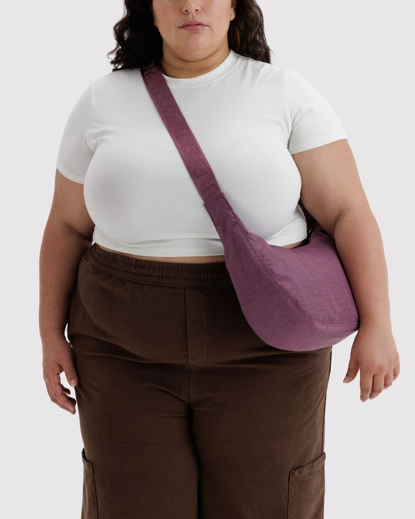 A model wearing a white t-shirt and brown pants, carrying a dusty purple fabric crossbody bag with a wide strap.
