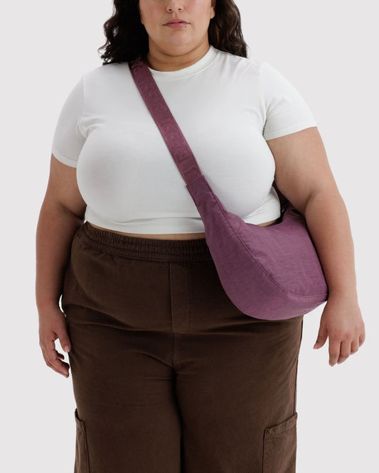 A model wearing a white t-shirt and brown pants, carrying a dusty purple fabric crossbody bag with a wide strap.