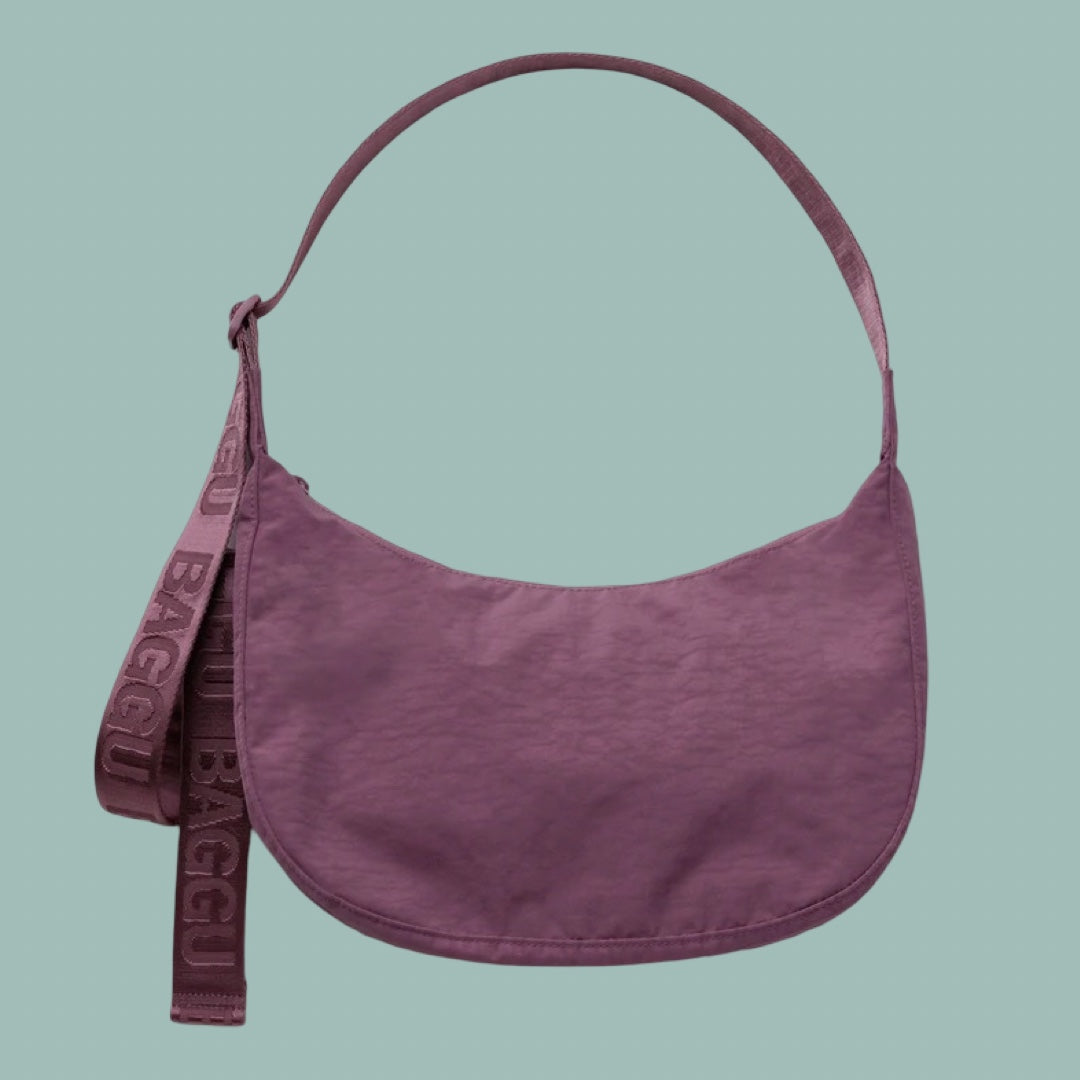 Baggu Medium Crescent Bag in Wild Plum. A dusty purple crescent-shaped shoulder bag with a matching wide strap featuring the embossed brand name BAGGU, set against a solid sage green background.