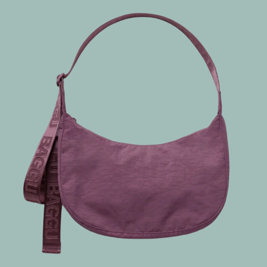 Baggu Medium Crescent Bag in Wild Plum. A dusty purple crescent-shaped shoulder bag with a matching wide strap featuring the embossed brand name BAGGU, set against a solid sage green background.