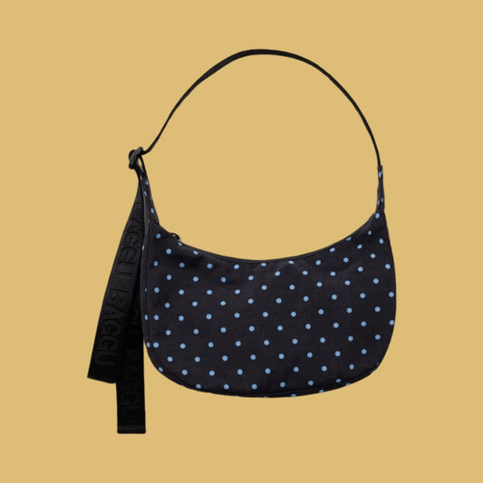 Baggu Small Crescent Bag - Black and Sky Blue Micro Dot. A crescent-shaped shoulder bag with a black base and light blue polka dots, featuring a black adjustable strap, set against a solid mustard yellow background.