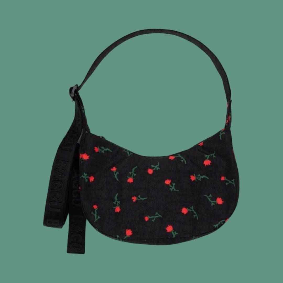 Baggu Small Crescent Bag in Painted Wildflower. A black crescent-shaped shoulder bag with a repeating red flower and green stem pattern, featuring a matching black strap, set against a solid teal background.