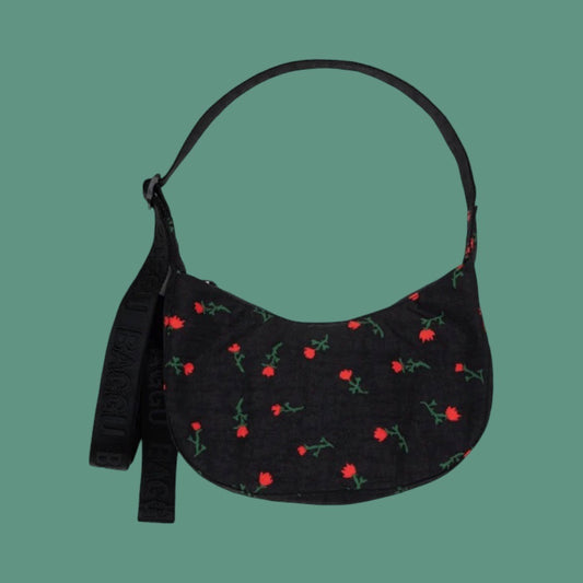 Baggu Small Crescent Bag in Painted Wildflower. A black crescent-shaped shoulder bag with a repeating red flower and green stem pattern, featuring a matching black strap, set against a solid teal background.