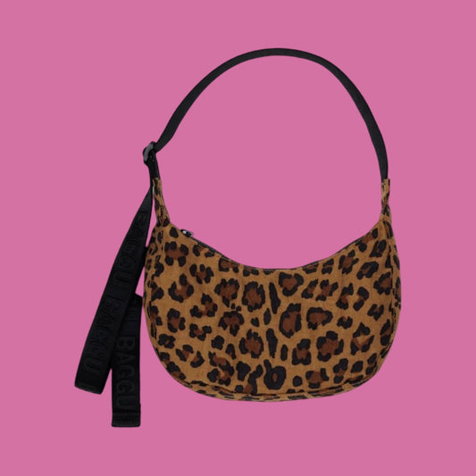 Baggu Small Crescent Bag - Leopard. A crescent-shaped shoulder bag with a brown and black leopard print pattern, featuring a black strap, set against a solid pink background.