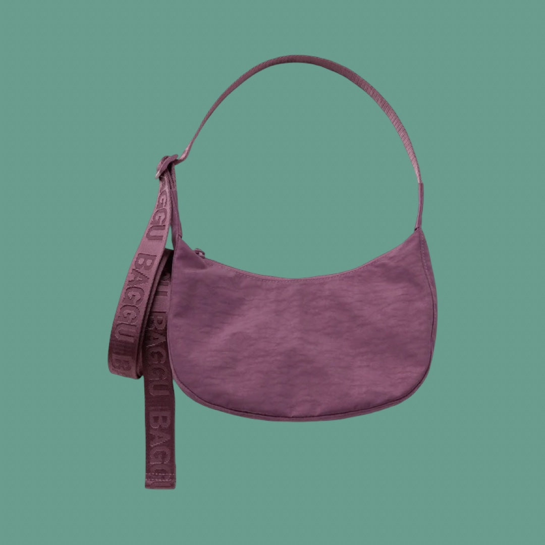 Baggu Small Crescent Bag in Plum. A dusty purple crescent-shaped shoulder bag with a matching wide strap featuring the brand name 'BAGGU' embossed, set against a solid teal-green background.