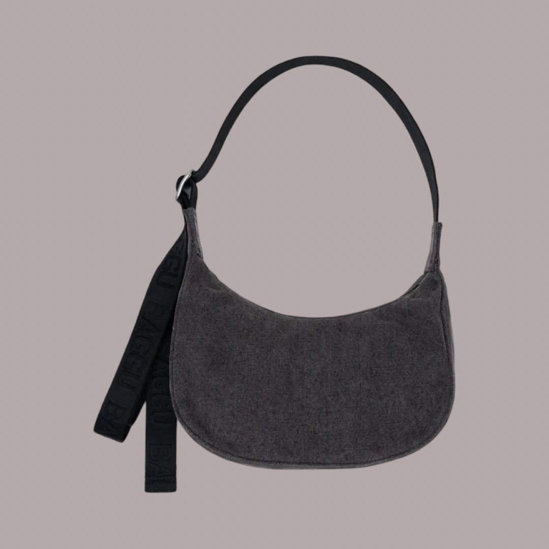 Baggu Small Denim Crescent Bag in Washed Black Denim. A dark gray denim crescent-shaped shoulder bag with a black strap, shown against a solid taupe-gray background.
